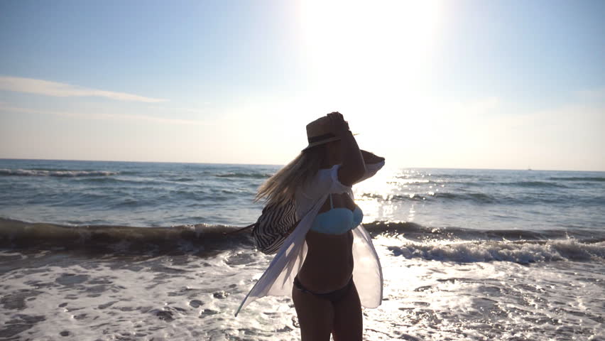 Young girl in bikini and shirt with backpack spinning on the beach near the sea. Beautiful woman in hat enjoying life and having fun at coast on a sunny day. Summer vacation or holiday. Slow motion | Shutterstock HD Video #1006709344