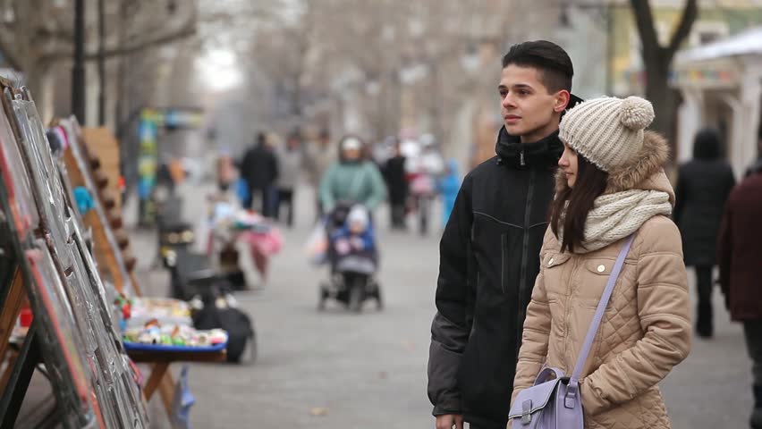 A romantic view of a happy young pair looking at stands with paintings on a park alley in winter.  A jolly girl smiles in a jolly way