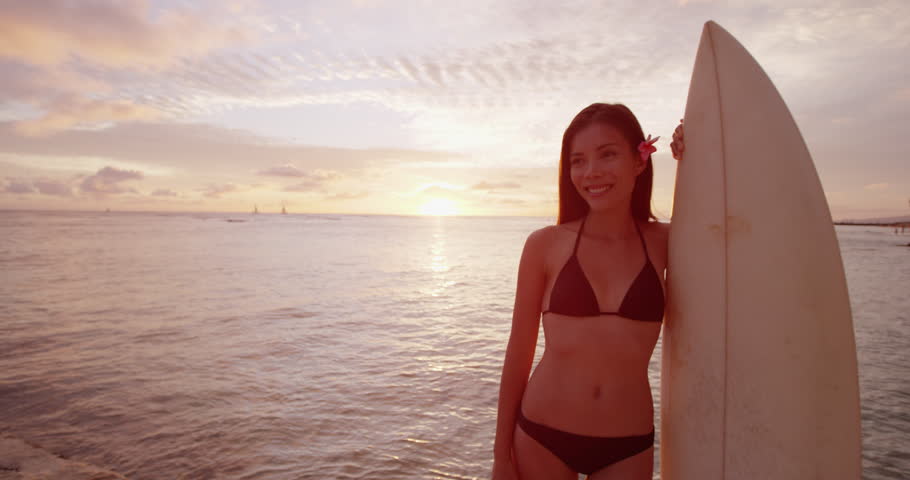 Young woman surfer going surfing - portrait of surfer girl standing with surfboard on beach at sunset. Multiracial model.