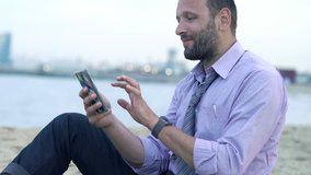 Young, happy businessman chatting on smartphone sitting on beach 
