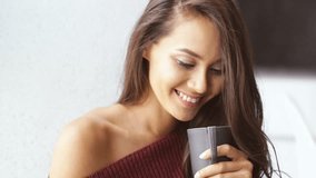 Close up view of Smiling brunette woman in sweater and jeans sitting near the window and using smartphone indoors