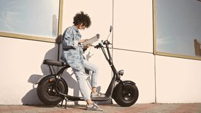 Side view of Smiling Brunette curly woman in sunglasses sitting on modern motorbike and using tablet computer outdoors