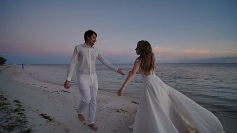 The bride and groom at sunset on a beautiful tropical beach. The bride sensually dances before the groom, holding on to the dress and turning. They are barefoot on the sandy shore of the ocean