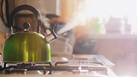 Boiling green kettle boiling with steam emitted from spout. The camera gently moves to the right. Solar glare from the kitchen window. Shallow depth of field
