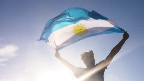 Young man holding argentinian national flag to the sky with two hands at the beach at sunset