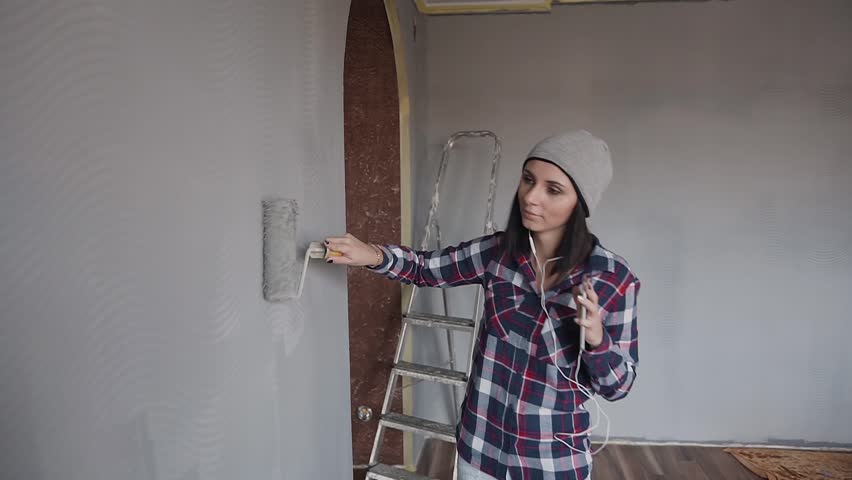 A charming young woman listens to music from the phone with headphones and finishes painting a part of a large wall with a gray paint and a brush roller. The hipsrer girl whitewash the walls at home | Shutterstock HD Video #1006831204