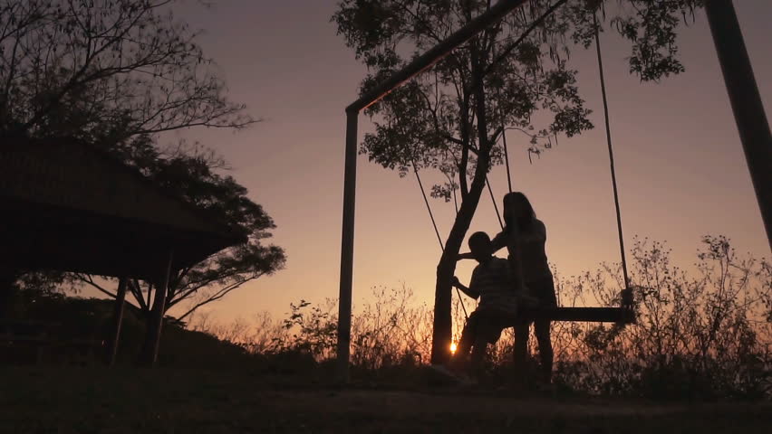 Slow motion,Silhouetted young mother with boy enjoying beautiful sunny day, swinging on swing set. Red orange sunset background.
