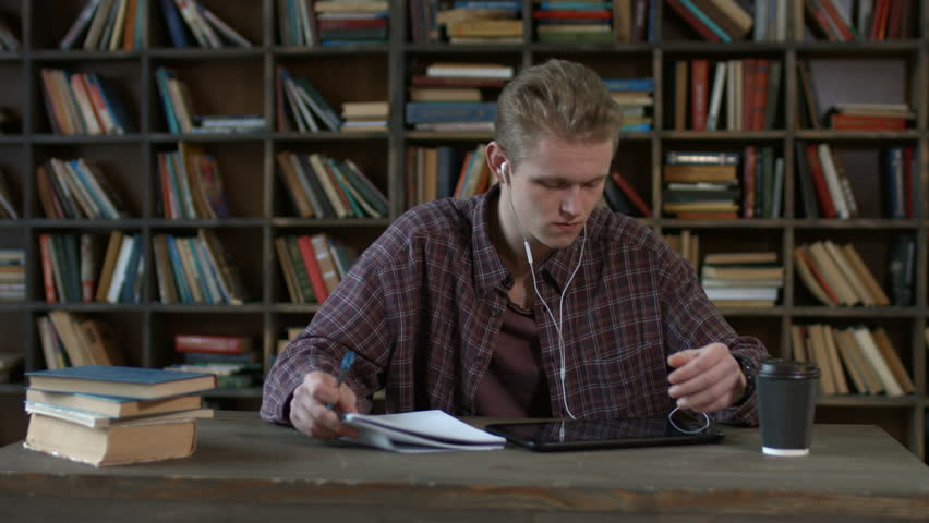Focused hipster student with digital tablet and headphones surfing the net and writing notes on textbook as sitting at the table in college library. Hadsome young man studying using tablet pc.
