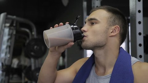 In the gym man with a towel around his neck drinks a protein cocktail. Young bodybuilder takes a sip from a bottle of vitamin drink to build muscle mass.