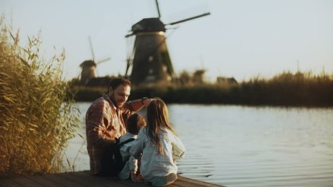 Single dad with two kids sit on a sunny lake pier. Traditional Dutch windmills in Netherlands. Family relationships. 4K.