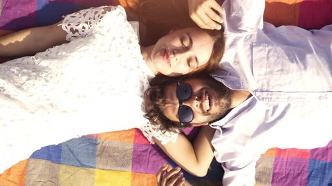 Happy lovely young couple lying on colorful blanket in the park looking at the sky pointing clouds romantic with guitar sunglasses beautiful attractive girl top view rotating camera slow motion