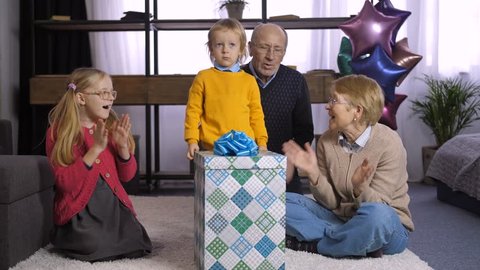 Cute little toddler boy unwrapping his birthday present from grandparents indoors. Sweet family clapping hands while grandson jumping from happiness, opening gift box and taking out his present