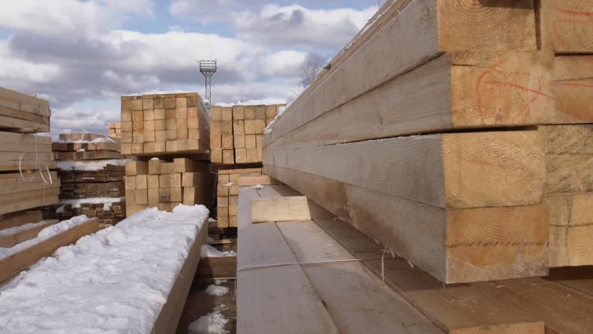 Piles of processed  packed wooden blocks stored in yard of sawmill, dirty ground with snow on sunny winter day
