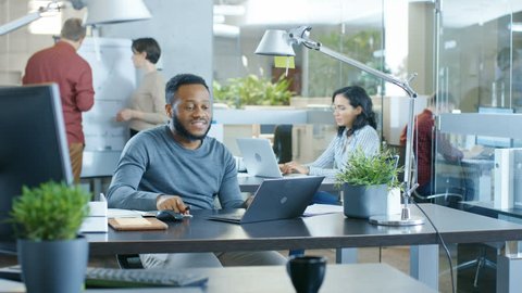 Black Man Working at His Desk Working on a Laptop, Encounters Funny Mistake and Laughs. In the Background Creative Young People Doing Their Jobs. Modern Bright Office. Shot on RED EPIC-W 8K Helium Cin