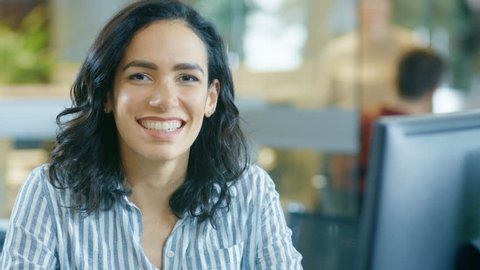 Portrait Shot of a Beautiful Young Hispanic Woman Working on a Computer, Smiling Warmly on the Camera. In the Background Busy Office with Working Colleagues. Shot on RED EPIC-W 8K Helium Cinema Camera