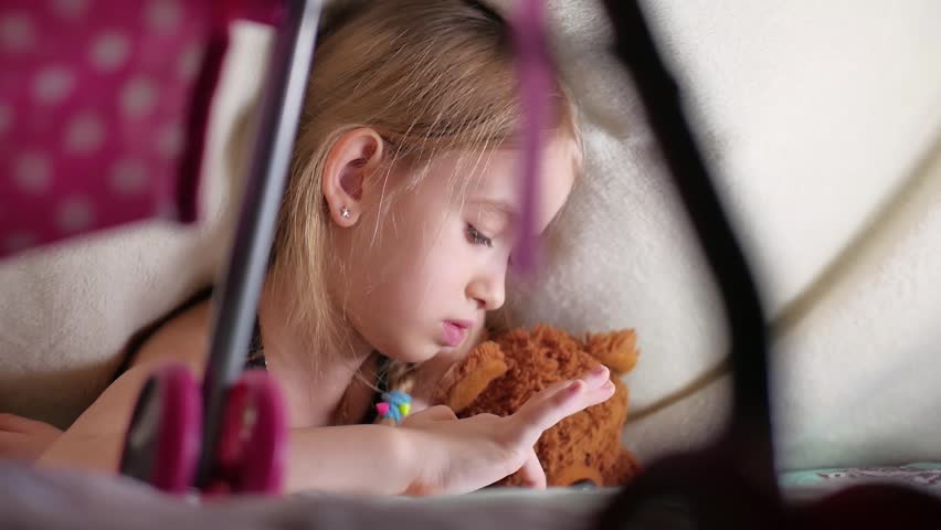 Cute little girl sitting on bed together with teddy bear playing games on tablet.