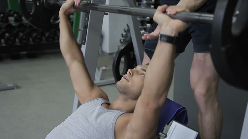 Young sportsman is doing his effort in inclined bench presses with his trainer on the gym. The man in grey sleeveless t-shirt is raising the barbell and his male couch in black sportswear is standing