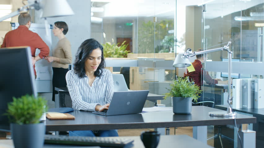 Busy International Office, Beautiful Hispanic Woman Working at Her Desk on a Laptop, She Writes Down information in Her Notebook. In the Background Her Coworkers. Shot on RED EPIC-W 8K Helium Camera.