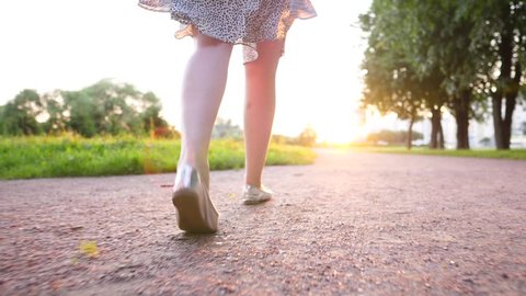 Female legs walk at gravel path, cinematic follow camera slide low behind. Fine evening sun shine ahead, vivid green colours of grass. Slender girl in summer dress have pleasure by stroll at city park