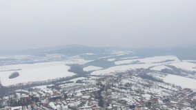 Video from drone of snowy, misty village or small town in winter time with hills on background. Bird perspective of village with forest.