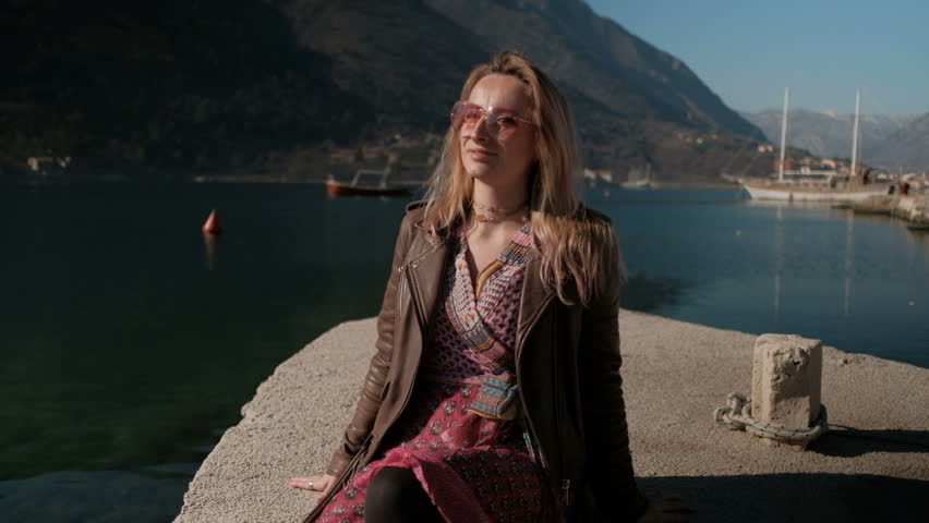 Young blond woman in pink glasses and dress sitting by the sea on a sunny day.
