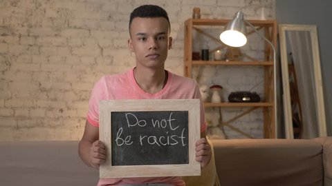 A sad African guy sits on the couch and holds a sign. Do not be racist. Anti-racism, national inequality concept. Look at the camera. 60 fps