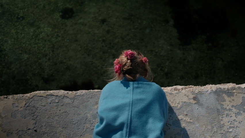 Top view of little blonde girl with roses in her hair and a blue coat lies on the waterfront and looks out to sea 