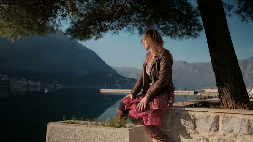 Young blond woman in pink glasses and dress sitting by the sea on a sunny day.