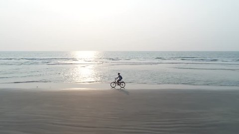 A young girl rides a Bicycle on the sandy beach against the sea and sunset. The shadow falls on the wet shore. Aerial view.