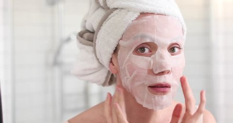 Woman applying cosmetic Japanese mask on her face and looking in the mirror looking at camera in the bathroom