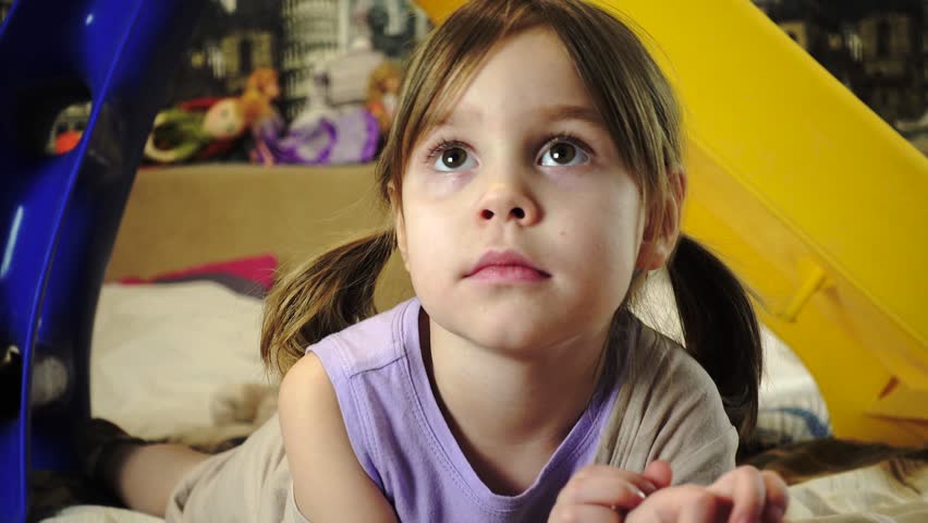 A girl with blonde hair and pigtails, of European or American appearance, is lying on the bed. To the child of 5-6 years. She is watching a movie. Close-up portrait.