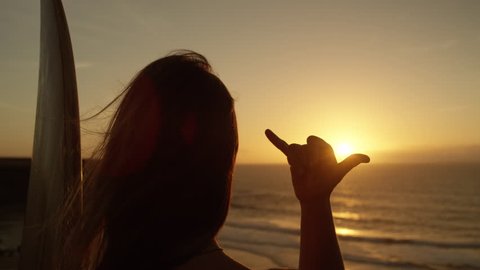 SLOW MOTION CLOSE UP: Unrecognizable girl with long flowing hair gazing at golden sunset giving the shaka sign. Female surfer making hang loose signal with her hand. Woman using Hawaiian hand gesture