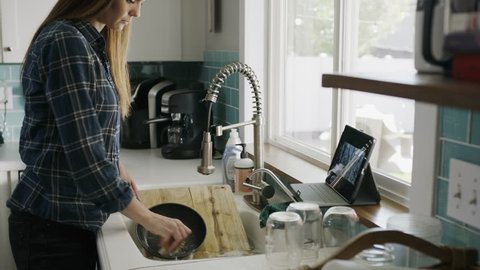 Woman washing dishes while watching streaming video on digital tablet / Alpine, Utah, United States