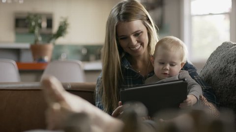 Panning shot of mother and baby daughter reading digital tablet in livingroom / Alpine, Utah, United States