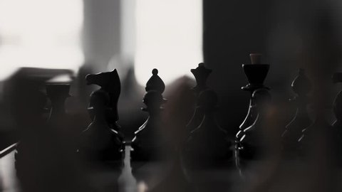 Part of white chess set standing on board in front of black chess set against window, close up