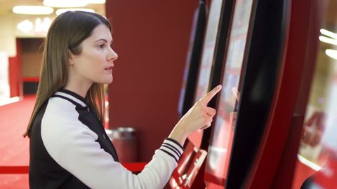 Teenager choosing movie and buying ticket from vending machine at movie theater at mall. Young woman making gestures by touching screen