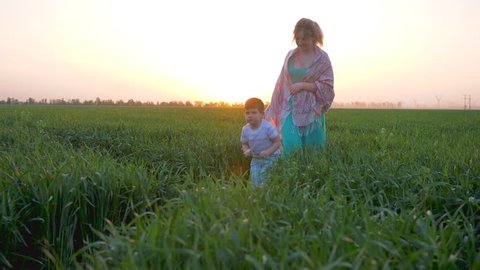 family holiday, young mother walks with joyful son through meadow in summer at sunrise in slow motion on against of pink heaven