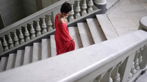 elegant actress in red dress on high heels climbs a large staircase at beautiful hall, top view