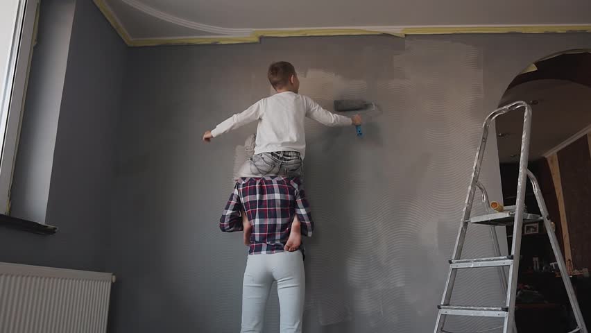 Young mother holds her son in her arms and the son paints a top wall with a roller in a gray color. Sister and brother draw wallpaper on the wall in gray color slow motion Royalty-Free Stock Footage #1007073799