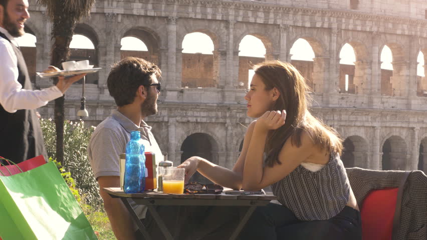 Elegant smiling waiter serving breakfast with coffee croissant cappuccino juice on a try outdoor to young happy couple in a bar restaurant table in front of colosseum in rome at sunset Royalty-Free Stock Footage #1007085619