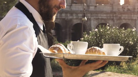Elegant waiter serving breakfast with breakfast coffee croissant cappuccino on a try outdoor to a bar restaurant table in front of colosseum in rome at sunset