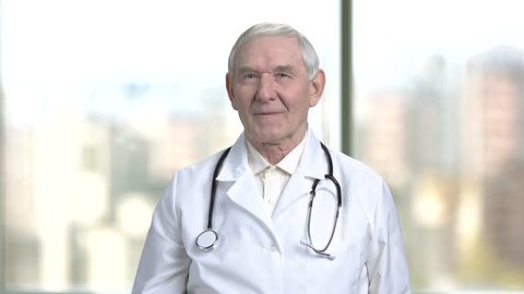 Happy old doctor shows thumb up. Senior physician with stethoscope shows you are fine. Blurred city from window background.
