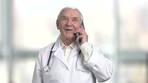 Positive cheerful senior doctor talking on the phone. Portrait of old physician with stethoscope talking on smartphone. Bright blurred windows background.