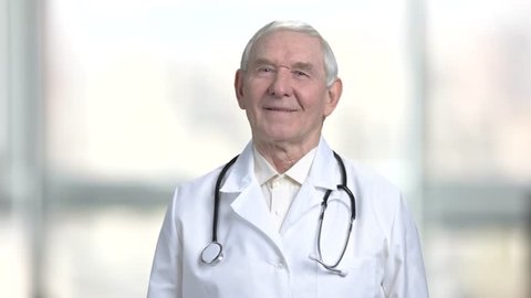 Portrait of senior doctor in abstract blurred bright background. Close up smiling old man in white coat and stethoscope.
