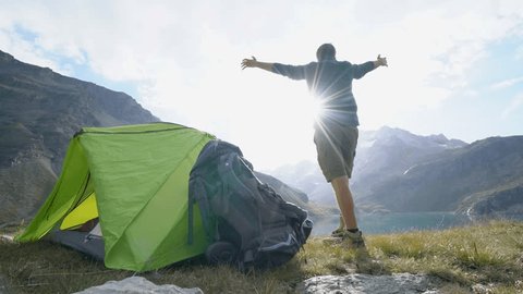 Young man on a hike stands by his tent near stunning mountain lake arms outstretched. Male hiker arms wide open in nature surrounded by scenic mountain lake landscape. Success at mountain top 