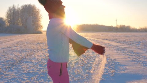 Playful young woman in a warm sweater, pink pants and red hat. Enjoying winter with snow outdoors on the field in warm sunrise at sunset. Throwing snow and standing under the snowfall. Slow motion, 4K : vidéo de stock