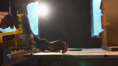 Side view shot of a professional carpenter using electric saw to deal with a wood boards. He is going along the drawn line.