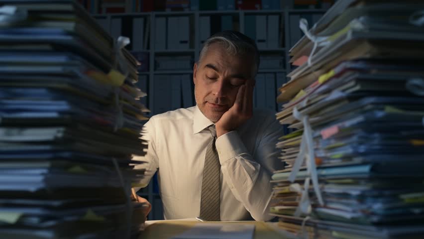 Tired businessman working late at night, he is overwhelmed by work and his desk is filled with paperwork, work overtime and deadlines concept | Shutterstock HD Video #1007116471
