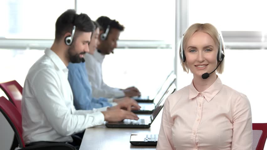 Blond woman with headset smiling in office. Lady smiling in call center office, office workers background. | Shutterstock HD Video #1007118019