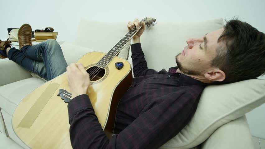 The young man lays on the couch and plays the guitar